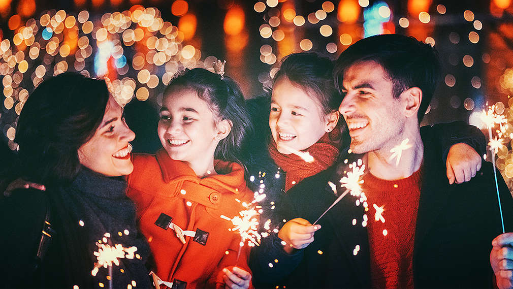 O familie îmbrăcată în haine de iarnă stă într-un loc luminat festiv, seara, afară, râzând și ținând în mâini artificii.