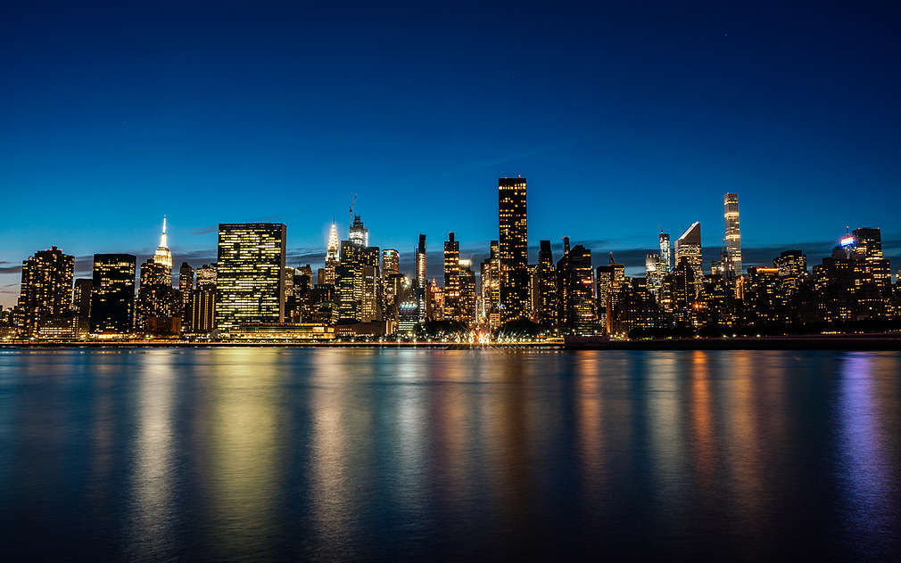 Widać odbijającą się w wodzie panoramę Nowego Jorku, w nocy, w niesamowitych odcieniach żółci, czerwieni, błękitu i fioletu.