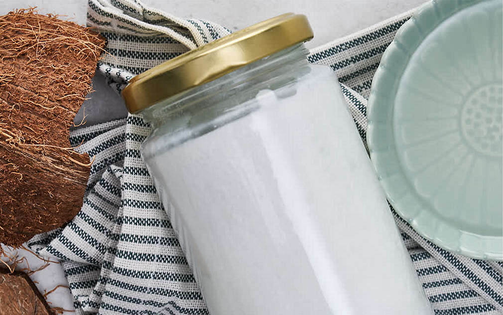 Ulei de cocos solid, într-un borcan închis, așezat pe un prosop de bucătărie cu dungi. Lângă acesta se poate vedea coaja unei nuci de cocos.