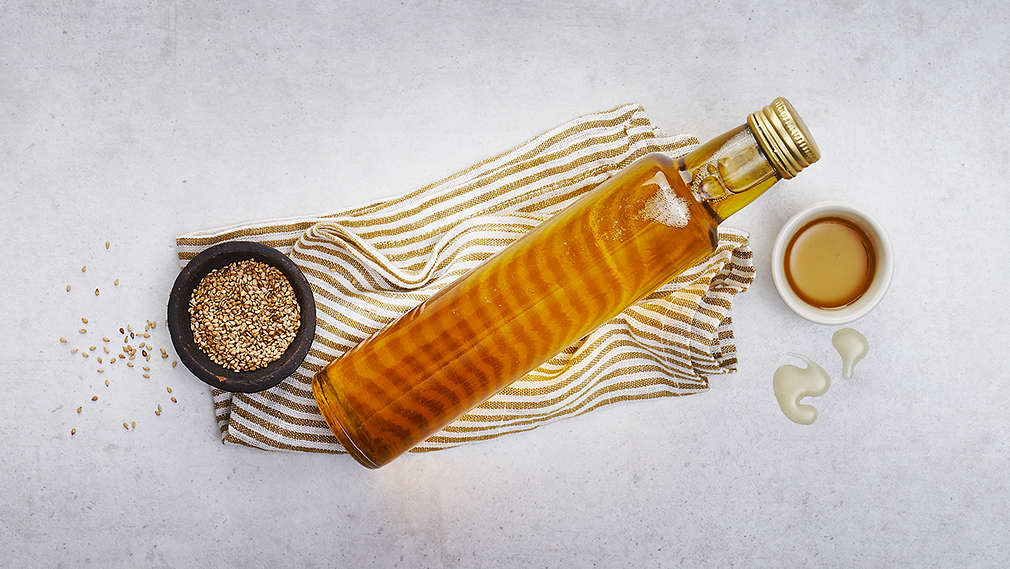 Вижда се купа със сусамови семена, друга купа със сусамово олио и бутилка сусамово олио, поставени върху раирана кухненска кърпа.