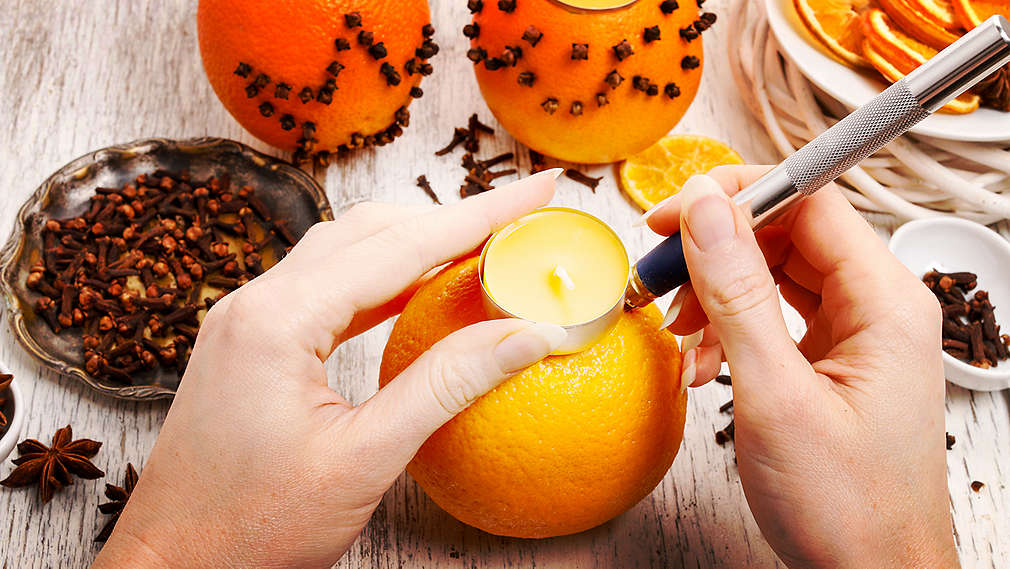 Dvije ženske ruke s pomoću noža izrađuju svijeću od naranče. Lijevo pored toga na stolu nalaze se klinčići.