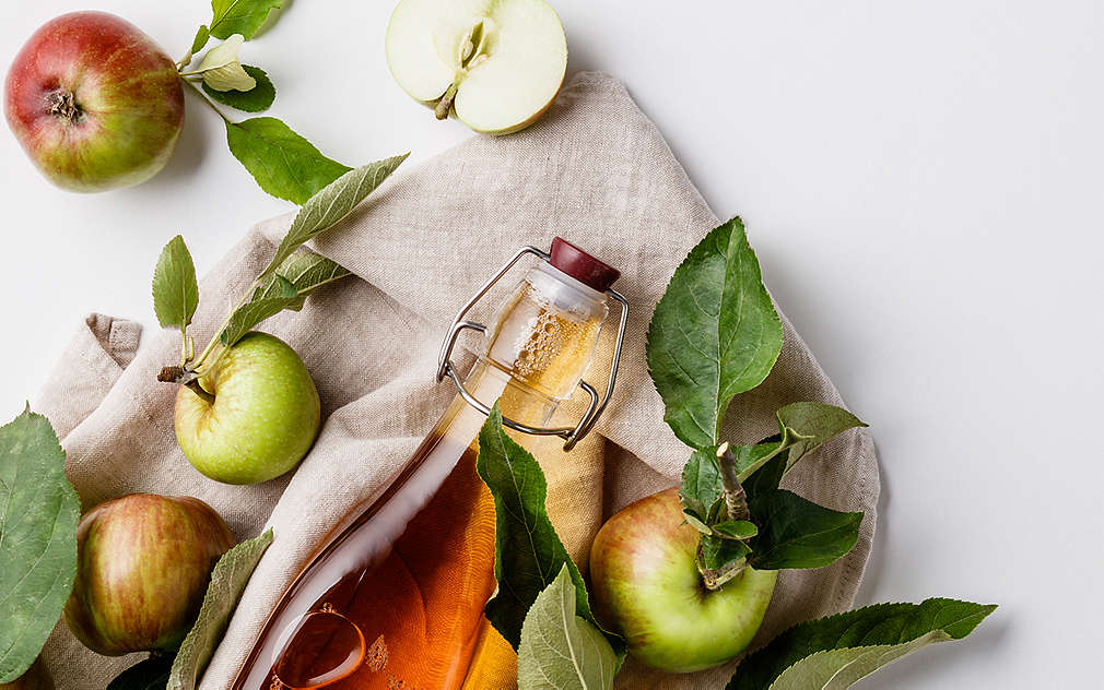 O sticlă de oțet de mere pe un prosop de bucătărie bej, alături de mere cu frunze.