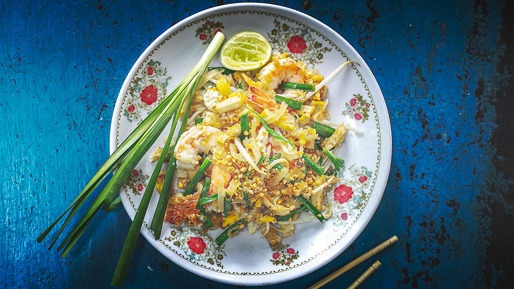 Dwie pałeczki i talerz świeżego pad thai z orzeszkami ziemnymi, krewetkami królewskimi, dymką i wyciśniętą limonką na niebieskim tle.