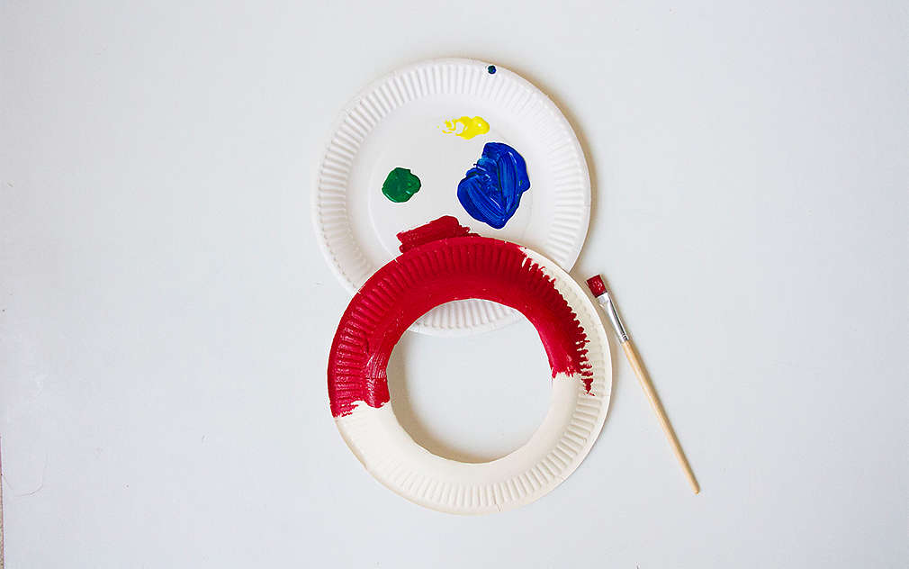 Un inel din farfurie de carton pictat cu roșu pe un fundal alb, pe o farfurie de carton cu pete de culoare, lângă care se află o pensulă.