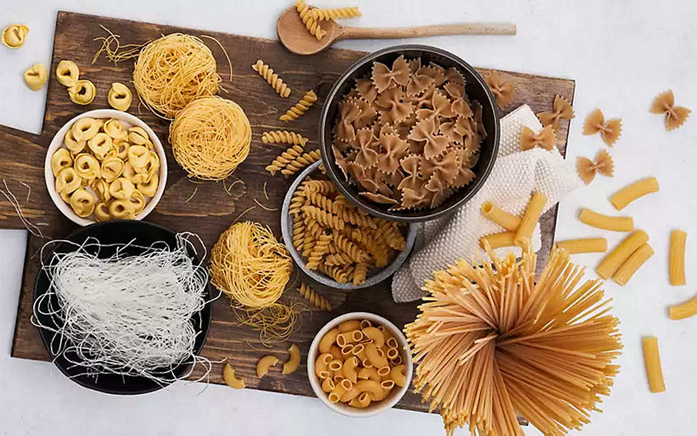 Različite vrste tjestenine nalaze se jedna do druge na drvenoj dasci ili u posudama. Daska se nalazi na svijetloj podlozi