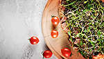 Se poate vedea o pinsa cu topping de salam, creson și germeni pe un tocător de lemn rotund, lângă câteva roșii cherry.