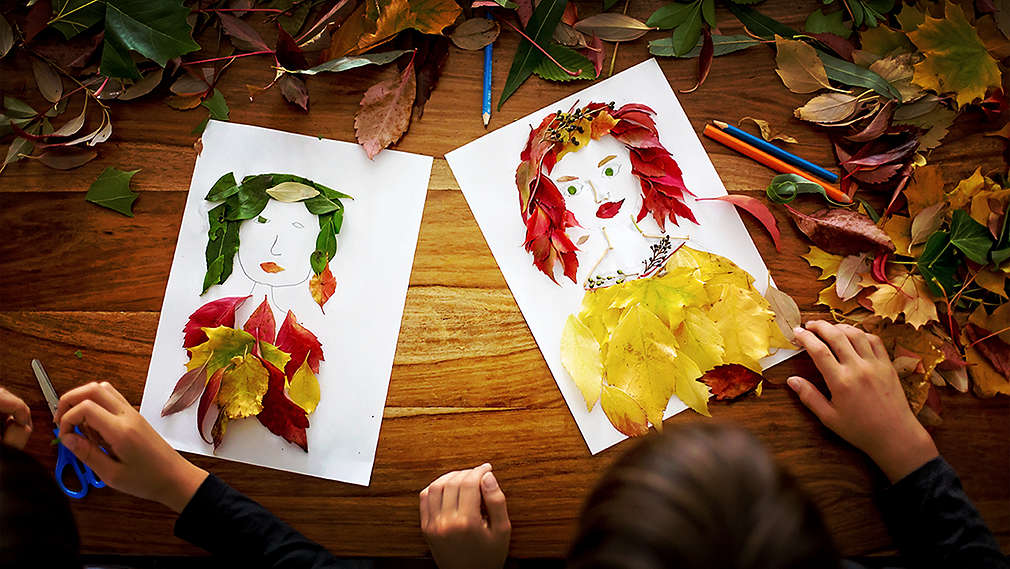 Doi copii, văzuți de sus, fac două portrete din frunze colorate la o masă de lemn pe care sunt așezate frunzele adunate și vopseaua.