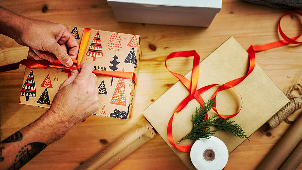 Na obrázku sú dve mužské ruky, ktoré balia vianočný darček. Na stole ležia okrem toho ďalšie pomôcky na balenie, ako napríklad baliaci papier, stuha a kartón.