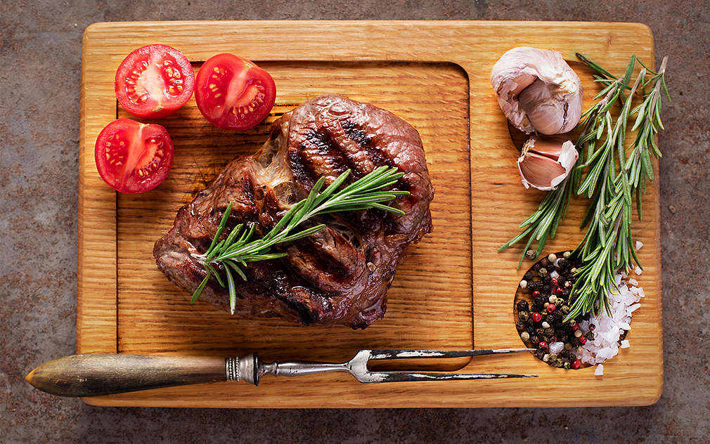 Готова пържола, розмарин и домати, поставени върху дървена кухненска дъска 