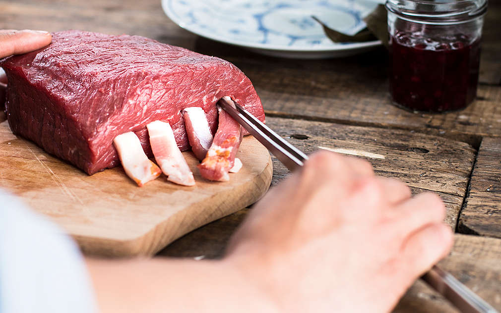 Widać dłoń przeciągającą paski boczku przez przekłutą porcję mięsa wołowego, które leży na drewnianej desce.