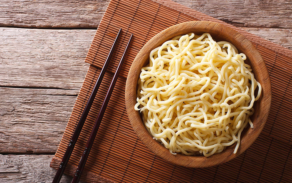 Tmavý drevený stôl, na ktorom je položené hnedé prestieranie z bambusu. Na ňom sú paličky a drevená miska naplnená uvarenými ramen rezancami.