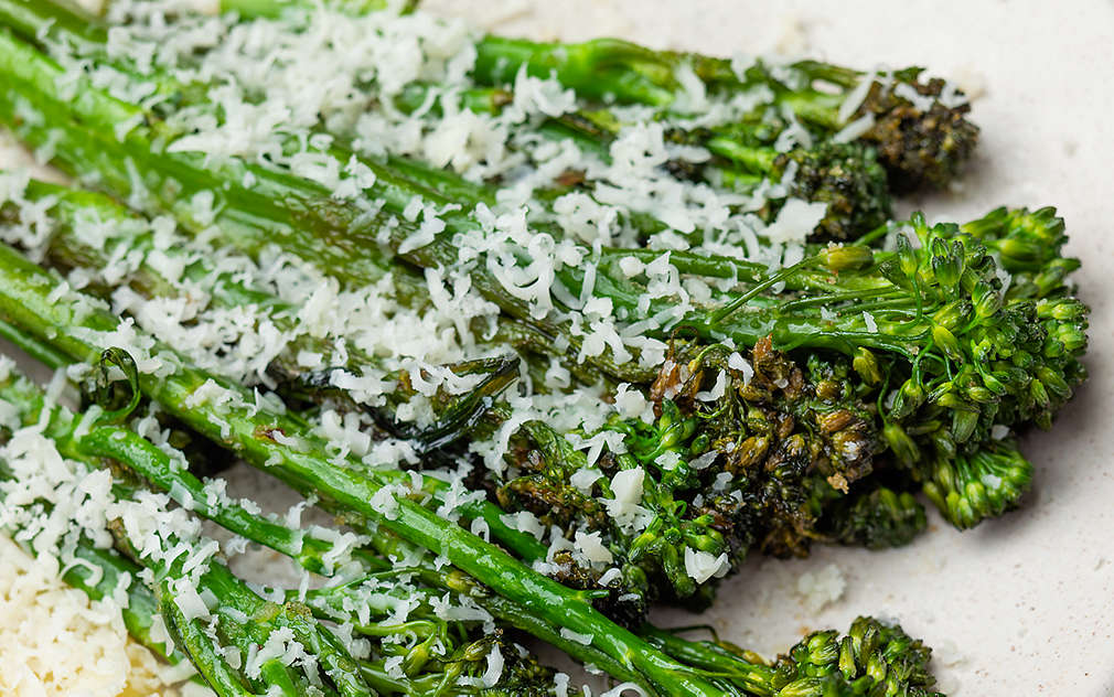 Pieczone bimi z plastrami cytryny i drobno startym parmezanem na głębokim białym talerzu ustawionym na białym tle.