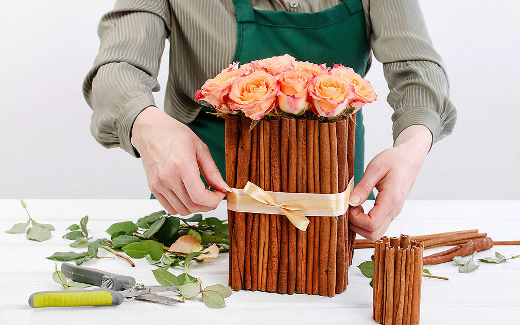 Suprafață albă, fundal gri deschis. O persoană cu un pulover verde deschis și un șort verde închis, doar cu trunchiul, brațele și mâinile vizibile, leagă o fundă în jurul unui recipient handmade din bucăți lungi de scorțișoară, în care sunt așezați trandafiri. De asemenea, pe suprafață mai sunt și un recipient mic cu bețe de scorțișoară, foarfece de grădină, tulpini și frunze de trandafir.