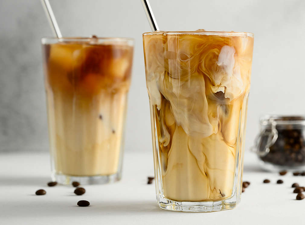 Ein Iced Coffee in einem Longdrink-Glas mit einem Strohhalm darin; daneben ein kleines Schälchen mit Kaffeebohnen