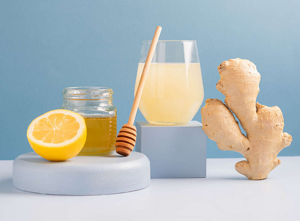 Ein Ingwer-Shot, eine Ingwerknolle, eine halbe Zitrone sowie ein Glas Honig mit einem Honiglöffel sind nebeneinander aufgereiht