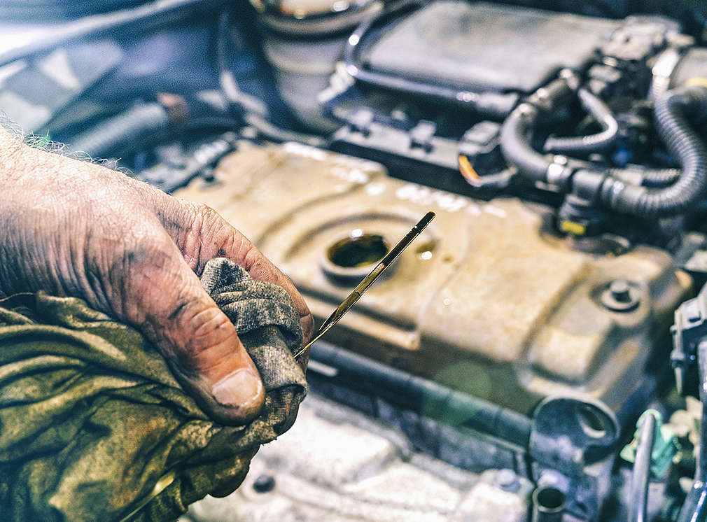 In einer Hand wird ein Ölstab gehalten. Im Hintergrund ist die Motorhaube zu sehen.