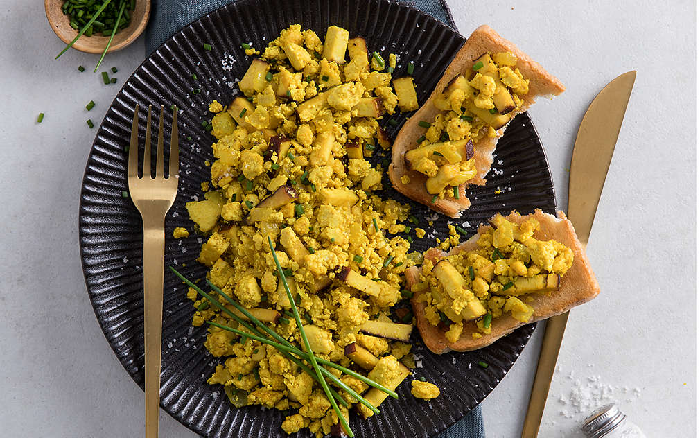 Goldenes Besteck, schwarzer Teller auf weißem Untergrund, links oben ein kleines Schälchen mit geschnittenem Schnittlauch. Auf dem Teller: Veganes Rührei aus Tofu mit Schnittlauch, zwei Toasthälften, belegt mit Rührei.