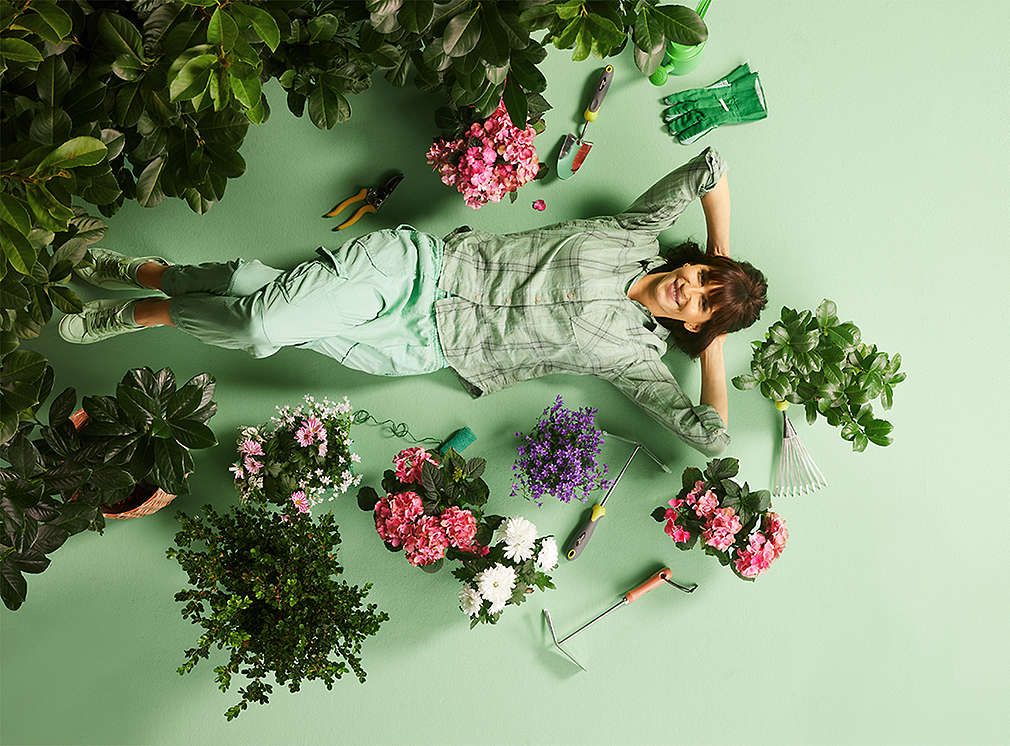 A woman lies on a green background surrounded by flowers and garden tools.