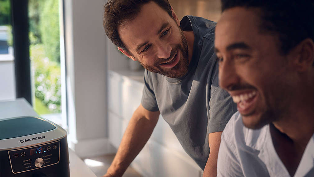 Zwei Männer unterhalten sich lachend in einer hellen Küche, während im Vordergrund das leuchtende Display einer SILVERCREST Heißluftfritteuse zu sehen ist., Zwei M�nner unterhalten sich lachend in einer hellen K�che, w�hrend im Vordergrund das leuchtende Display einer SILVERCREST Hei�luftfritteuse zu sehen ist.