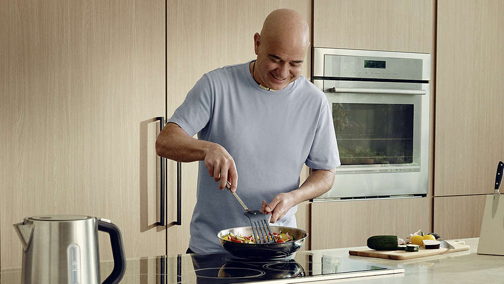Andre Agassi steht lächelnd in einer modernen Holzküche an einem Induktionskochfeld und wendet mit einem Pfannenwender frisches Gemüse in einer Edelstahlpfanne., Andre Agassi steht l�chelnd in einer modernen Holzk�che an einem Induktionskochfeld und wendet mit einem Pfannenwender frisches Gem�se in einer Edelstahlpfanne.
