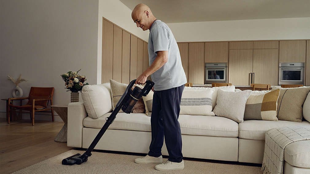 Andre Agassi nutzt einen kabellosen SILVERCREST Staubsauger, um den hellen Teppich in einem modern eingerichteten Wohnzimmer vor einer hellen Couchgarnitur zu saugen.