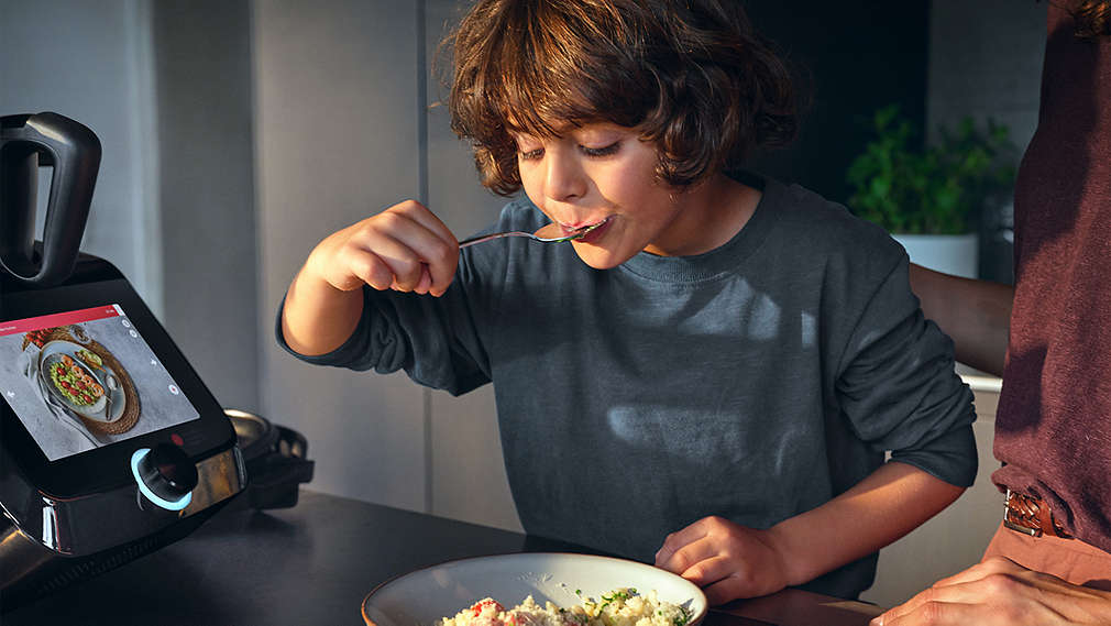 Ein Junge mit braunem Lockenkopf isst genüsslich von einem Teller, während im Vordergrund das Display einer SILVERCREST Monsieur Cuisine ein Rezeptbild anzeigt., Ein Junge mit braunem Lockenkopf isst gen�sslich von einem Teller, w�hrend im Vordergrund das Display einer SILVERCREST Monsieur Cuisine ein Rezeptbild anzeigt.