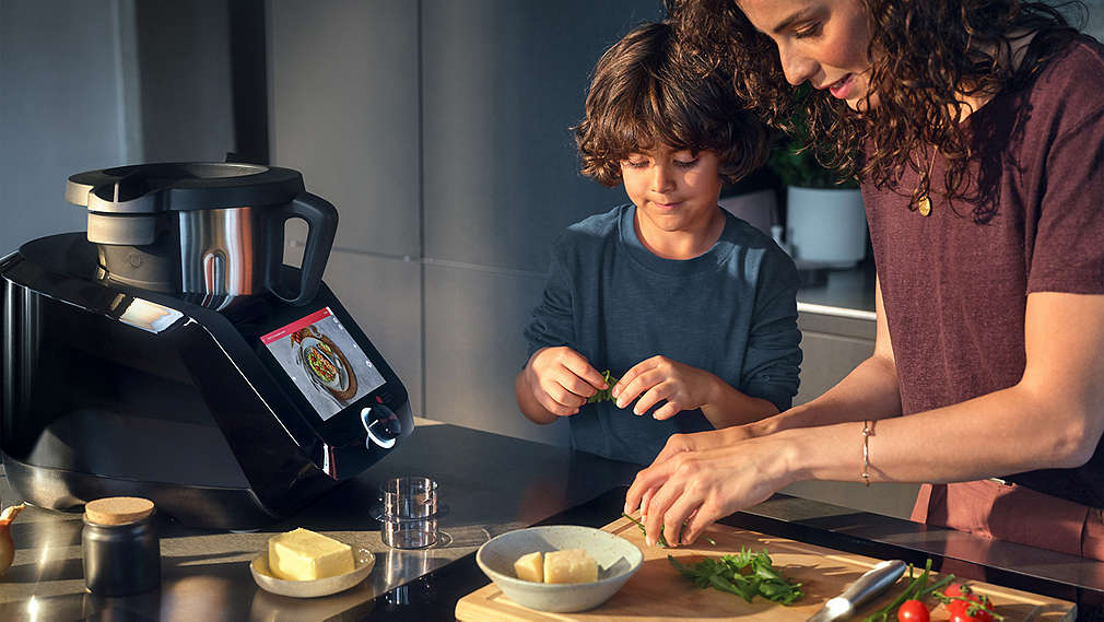 Eine Frau und ein Junge schneiden gemeinsam frische Kräuter auf einem Holzbrett, während daneben die SILVERCREST Monsieur Cuisine Küchenmaschine betriebsbereit auf der Arbeitsplatte steht., Eine Frau und ein Junge schneiden gemeinsam frische Kr�uter auf einem Holzbrett, w�hrend daneben die SILVERCREST Monsieur Cuisine K�chenmaschine betriebsbereit auf der Arbeitsplatte steht.