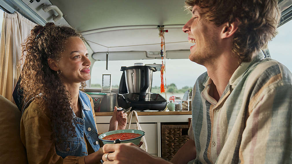 Ein lachendes Paar sitzt gemütlich in einem ausgebauten Campervan und isst aus Schalen, während im Hintergrund auf der Küchenzeile eine SILVERCREST Monsieur Cuisine steht., Ein lachendes Paar sitzt gem�tlich in einem ausgebauten Campervan und isst aus Schalen, w�hrend im Hintergrund auf der K�chenzeile eine SILVERCREST Monsieur Cuisine steht.