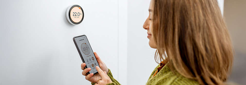 A person operates a modern, round digital thermostat on the wall and uses a smartphone app to set the temperature.