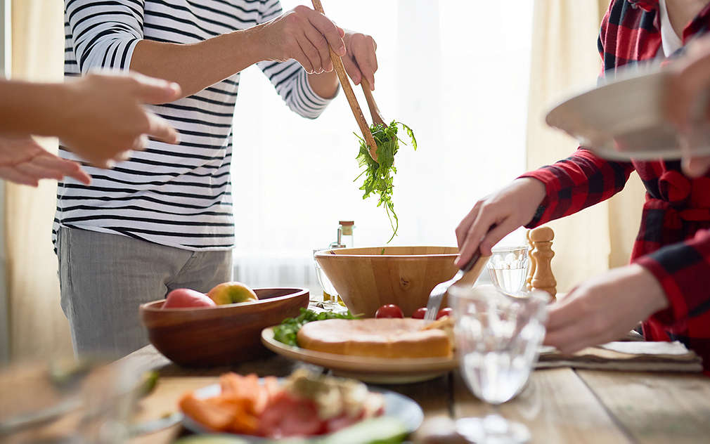 Trei persoane la masă, care se servesc cu salată. Pe masă există un bol cu mere, un bol cu salată, farfurii și pahare.