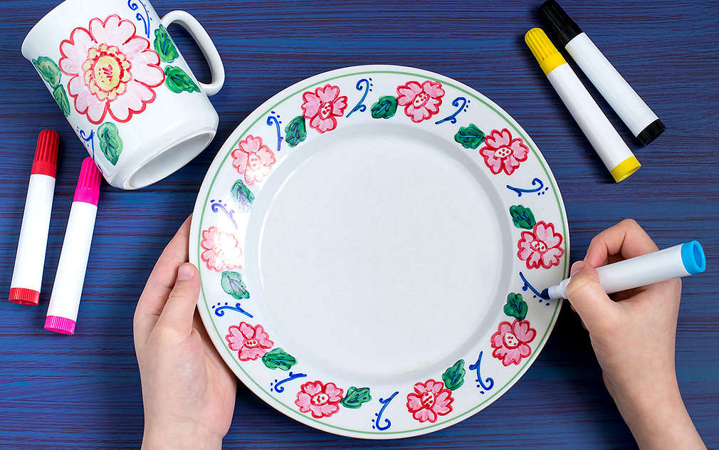 Tmavomodré pozadie, na ňom fixky v rôznych farbách, biela pomaľovaná keramická šálka, dve ruky a jeden tanier. Ruky držia a zdobia biely tanier fixkou. Okraj taniera je zdobený kvetmi.