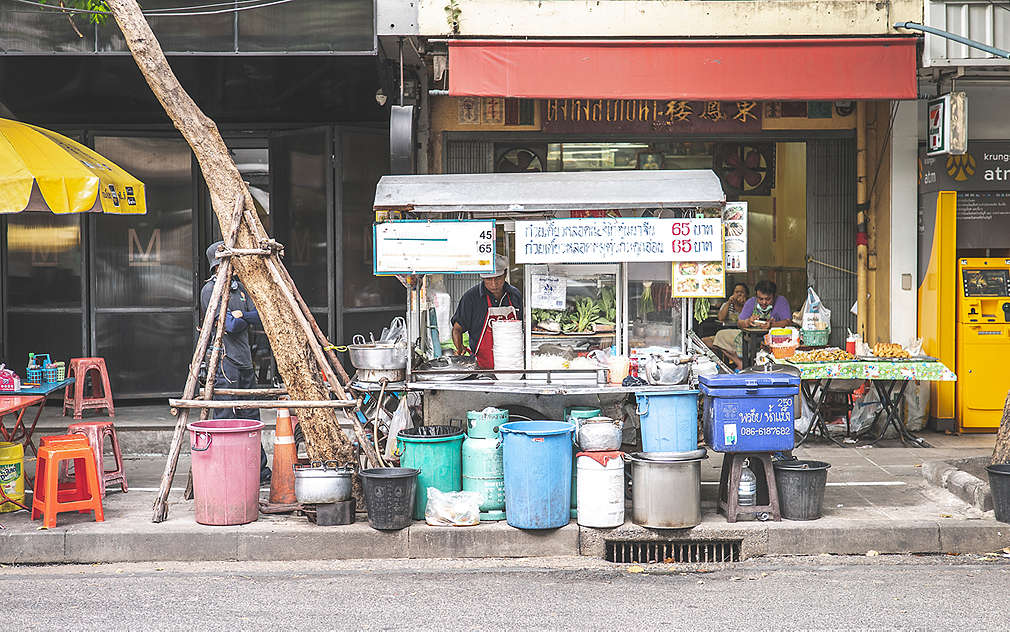 Na obrázku je typická pojízdná kuchyně v ulicích Bangkoku, kde se na pánvi wok připravuje čerstvé pad thai a prodává se jako street food. Vedle stánku s občerstvením je žlutý slunečník se stolkem a stoličkami na sezení.
