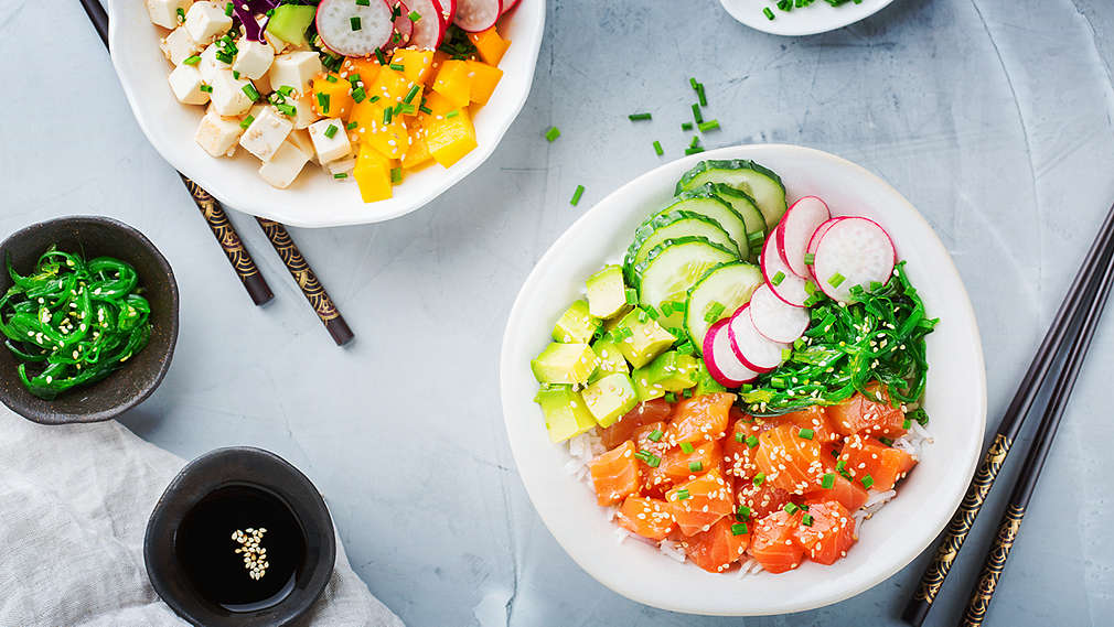 Se poate vedea un sushi bowl, felii de somon crud, felii de avocado, castraveți, ridichi și alge. În stânga, se poate vedea un alt bol cu orez, felii de tofu, mango, ceapă roșie, avocado și ridichi. De asemenea, în imagine: două seturi de bețișoare și trei boluri cu ceapă verde tocată, alge și sos de soia.