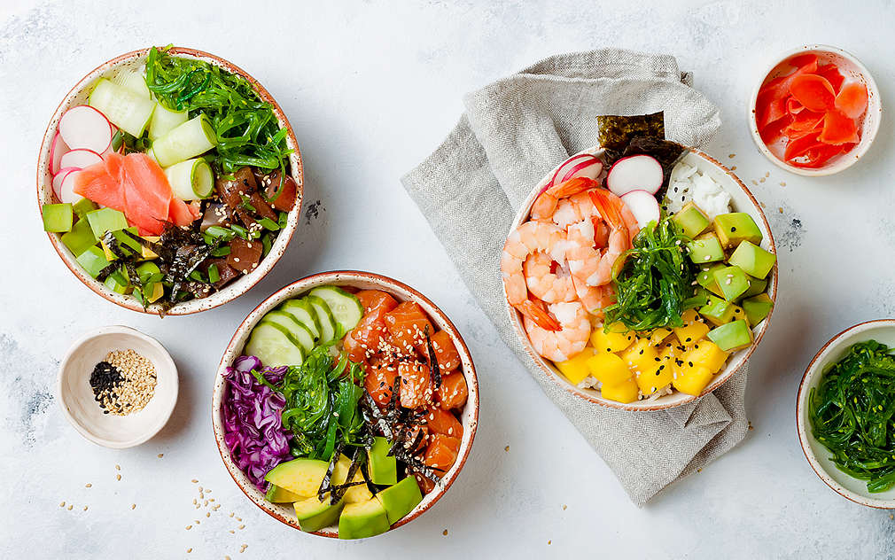 Tri zdjele s različitim sadržajem, uključujući tunu, škampe, avokado, krastavac, crveni luk, rotkvice, alge. Također na stolu: siva krpa za suđe, zdjelice sa sezamom, kimchijem i algama.