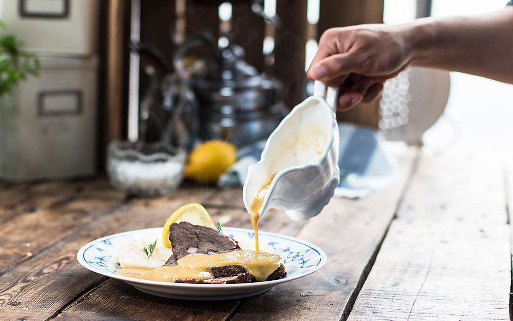 Widać dłoń polewającą sosem porcję svíčkovej z czeskimi knedlikami na talerzu na drewnianym stole.
