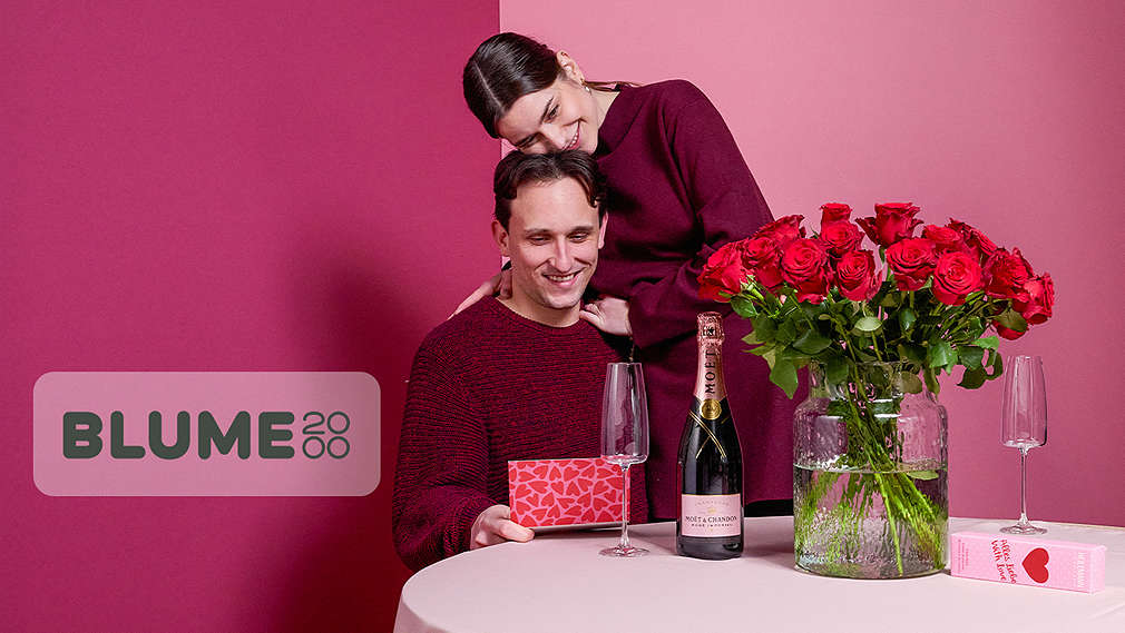 Abbildung: Ein Mann und eine Frau am Tisch mit einem Blumenstrauß, einer Flasche Sekt und einer Karte; Logo: BLUME2000