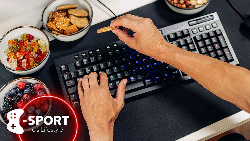 Eine Person greift über eine Gaming-Tastatur hinweg nach einem Cracker, daneben eine Gaming-Maus sowie Schälchen mit Beeren und Gummibärchen