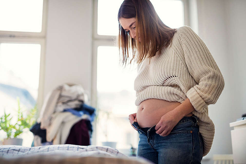 Изображение на бременна жена, която се опитва да си обуе вече омалели дънки