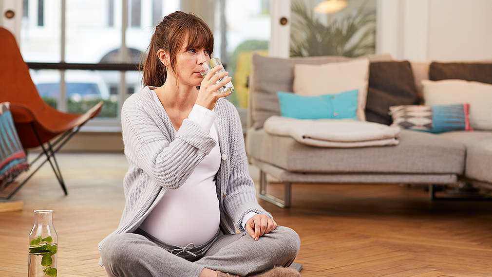 Schwangere sitzt im Schneidersitz und trinkt ein Glas Wasser