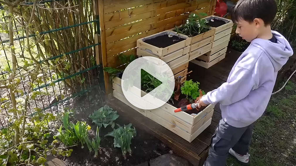 Junge hält vor einem Hochbeet einen Gartenschlauch in der Hand