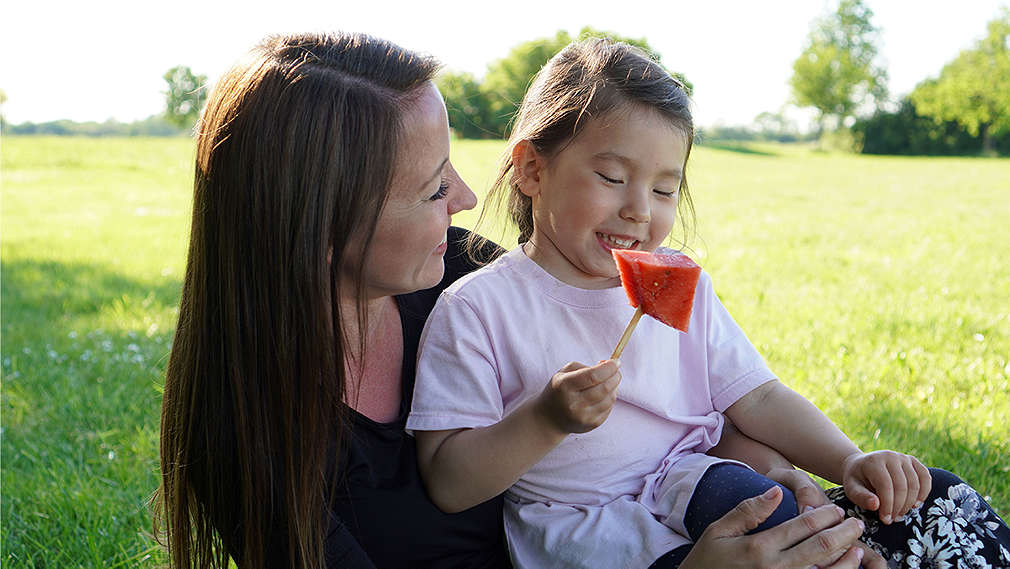 Mutter und Tochter sitzen auf einer Picknickdecke und essen Melone am Stiel