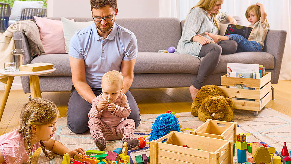 Zwei Kinder spielen gemeinsam und Vater sitzt daneben. Ein weiteres Kind sitzt mit Mutter auf dem Sofa.
