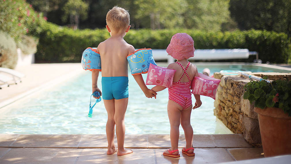 Ein Mädchen in rosaferbenem Badeanzug und ein Junge in blauer Badeshort stehen an einem Pool