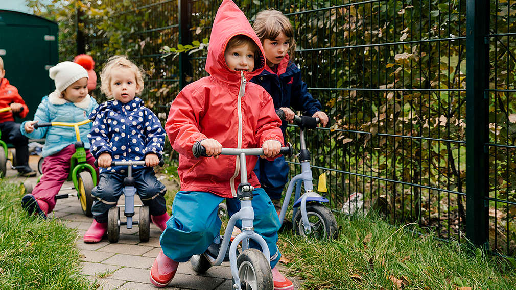 Kinder in Regenkleidung fahren auf einem schmalen Weg hintereinander Tretrad