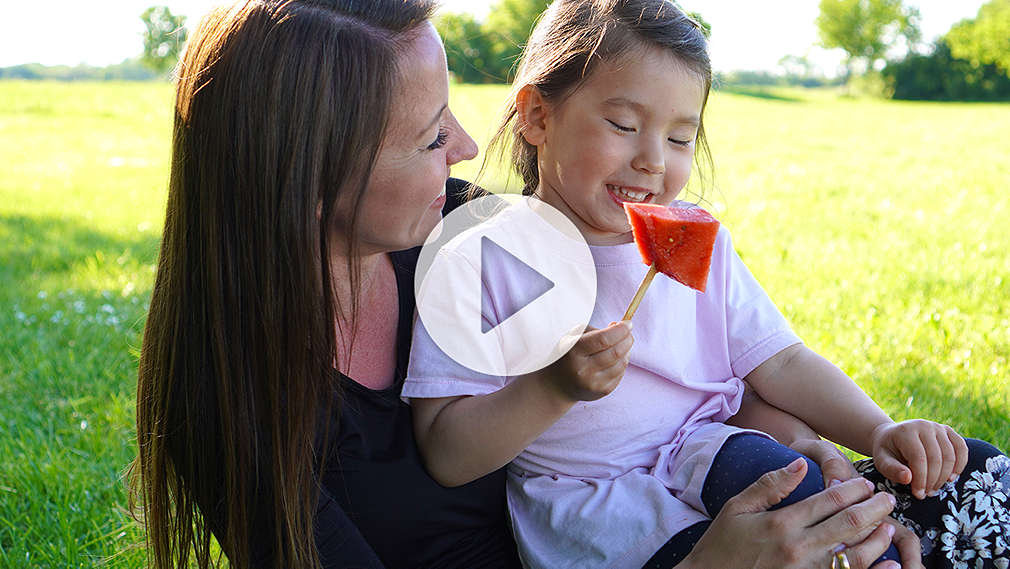 Mutter und Tochter sitzen auf einer Picknickdecke und essen Melone am Stiel