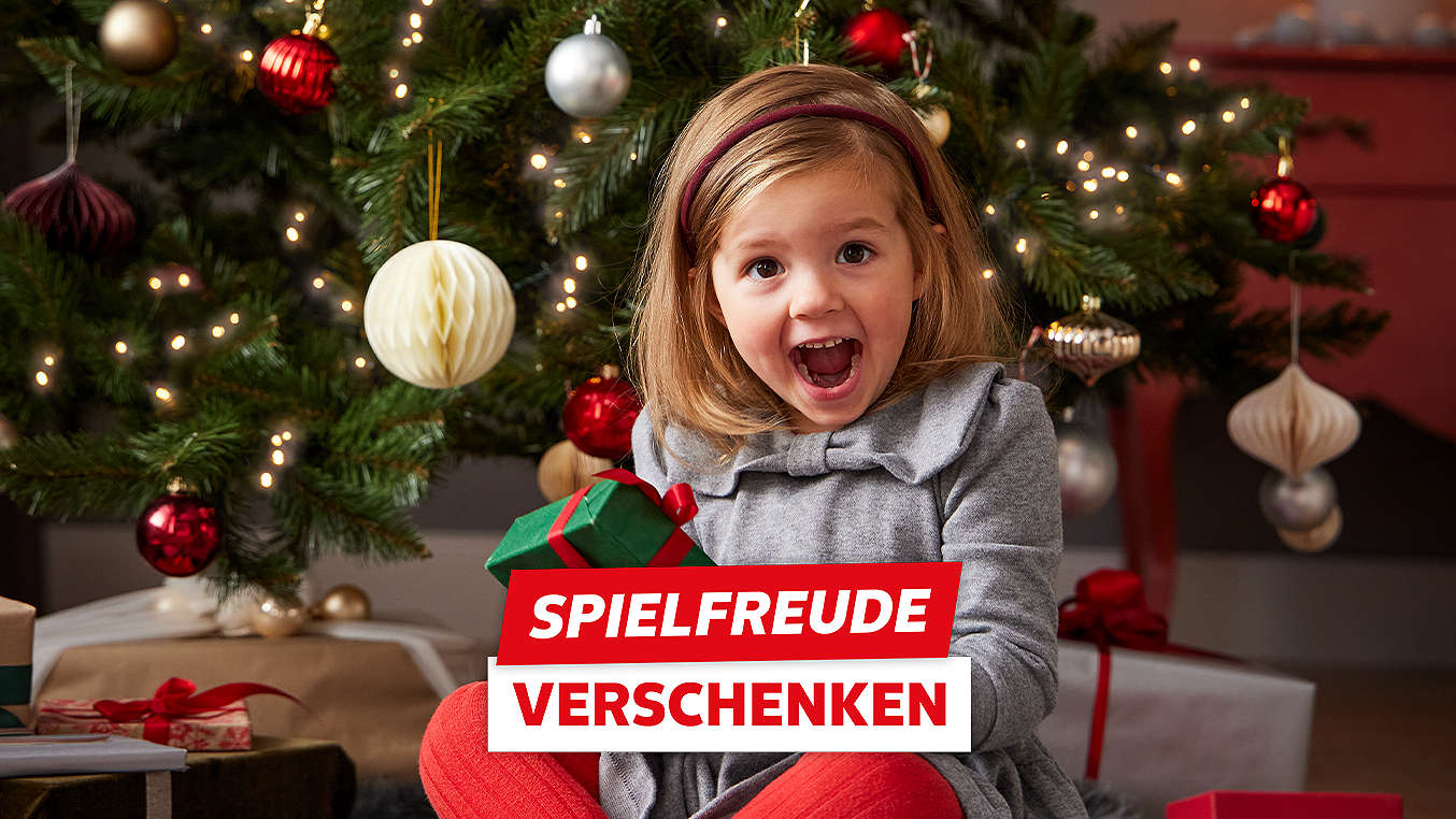 Schriftzug: Spielfreude verschenken; Abbildung: Kind unter einem Tannenbaum mit Geschenk in der Hand