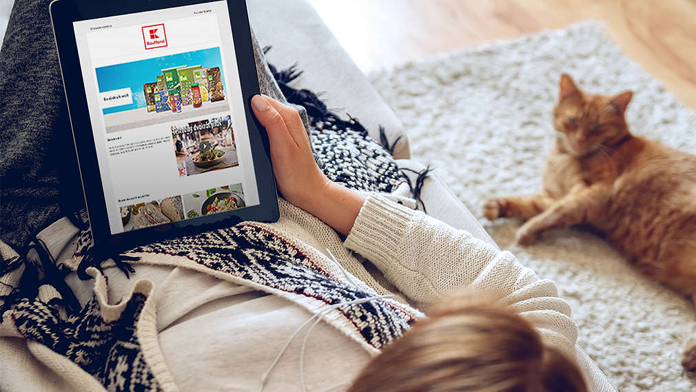 Žena si listuje akčním letákem Kaufland na tabletu a vedle ní leží kočka