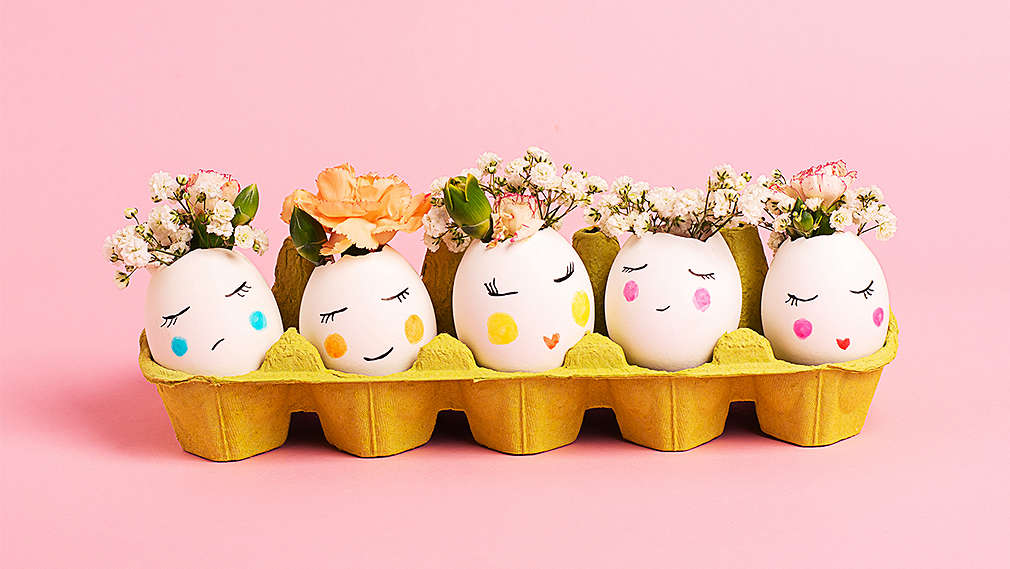 Hintergrund und Untergrund rosa, darauf ein 10er-Eierkarton und darin ausgeblasene weiße Eier mit kleiner Öffnung, in denen kleine Blumensträußchen sind. Die Eier haben Gesichter mit geschlossenen Augen, leuchtenden Wangen in unterschiedlichen Farben und kleine Münder.