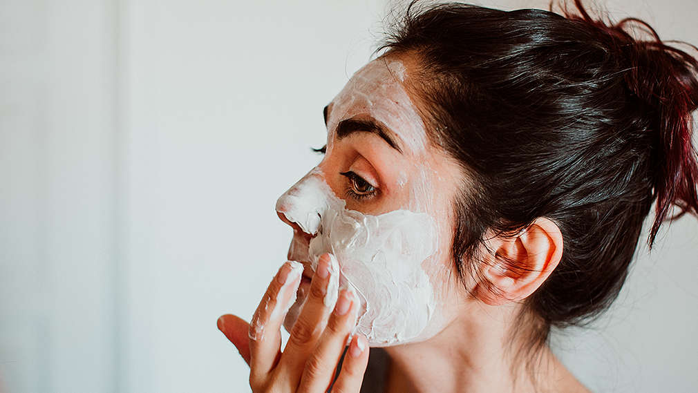 Eine junge Frau mit zusammengebundenen Haaren in ihrem Badezimmer, die eine cremige Maske auf ihr Gesicht aufträgt 