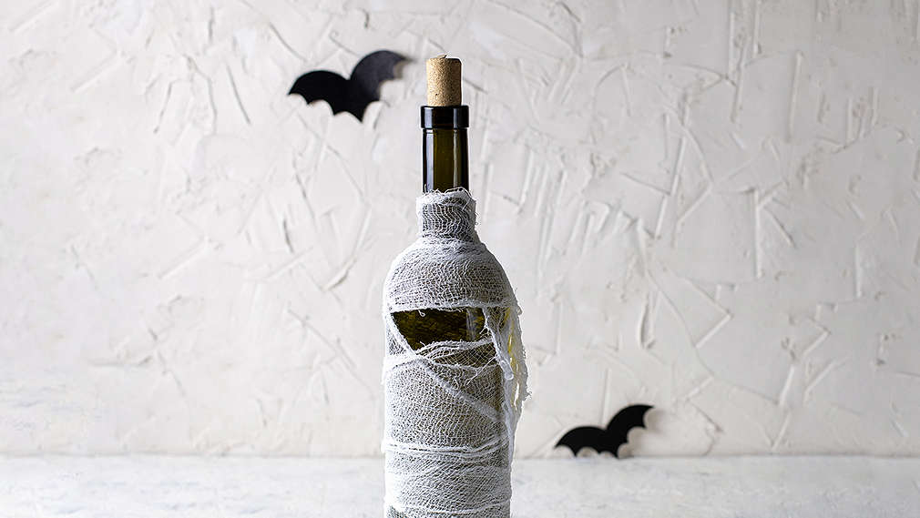 Weiße Wand mit Struktur im Hintergrund, hellgrauer Untergrund, darauf eine Weinflasche mit Korken, die mit Mullbinde umwickelt ist. Als Dekoelemente sind außerdem drei kleine Fledermäuse aus Papier im Bild.